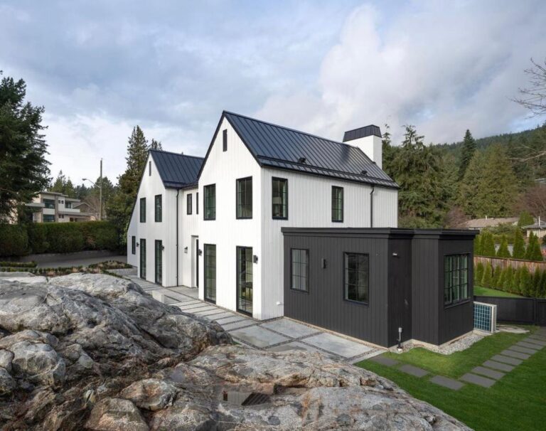 White board-and-batten modern farmhouse with a black standing-seam metal roof and black-clad addition on a rocky, treed lot